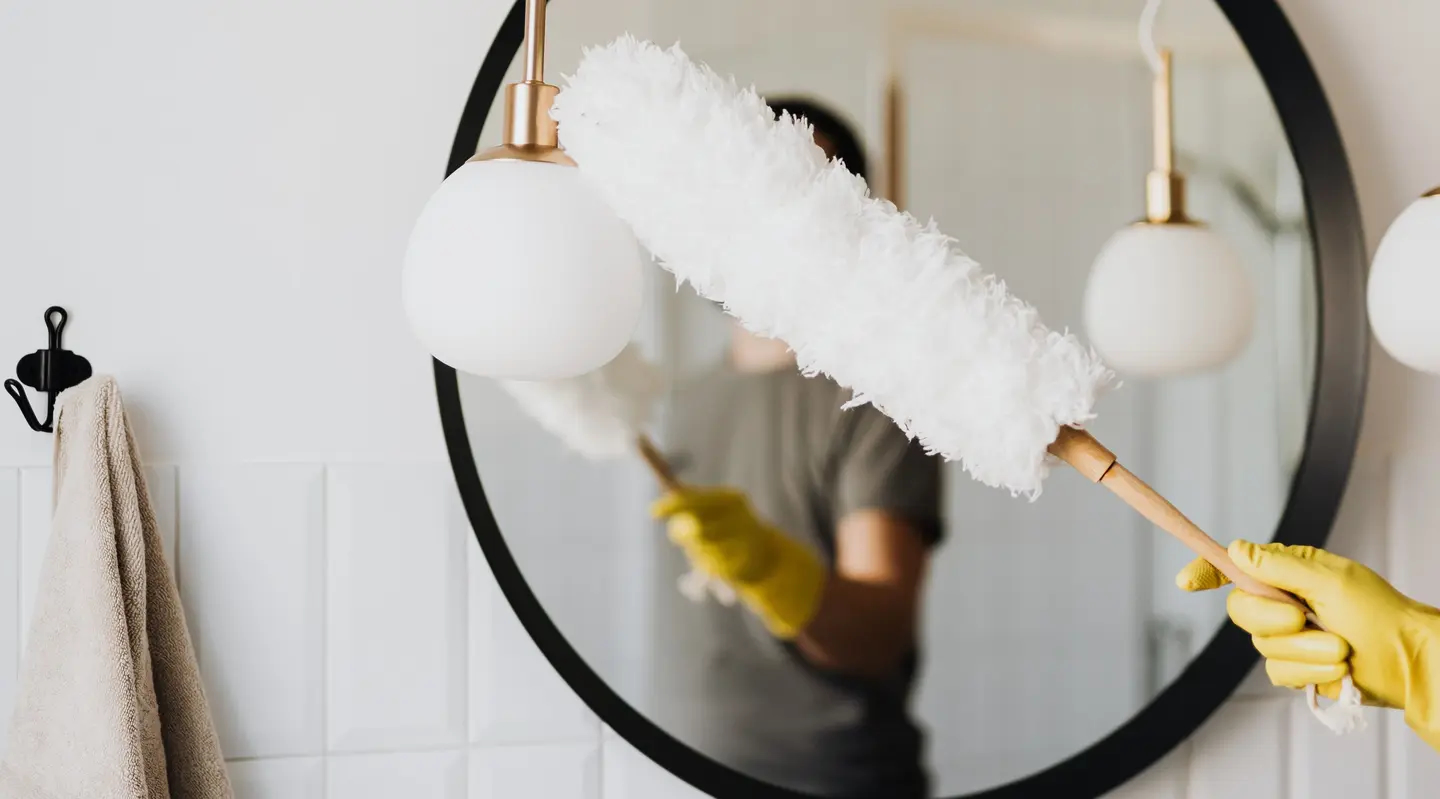 Person dusting a round mirror with a white duster, wearing yellow gloves. A towel hangs on a nearby hook.