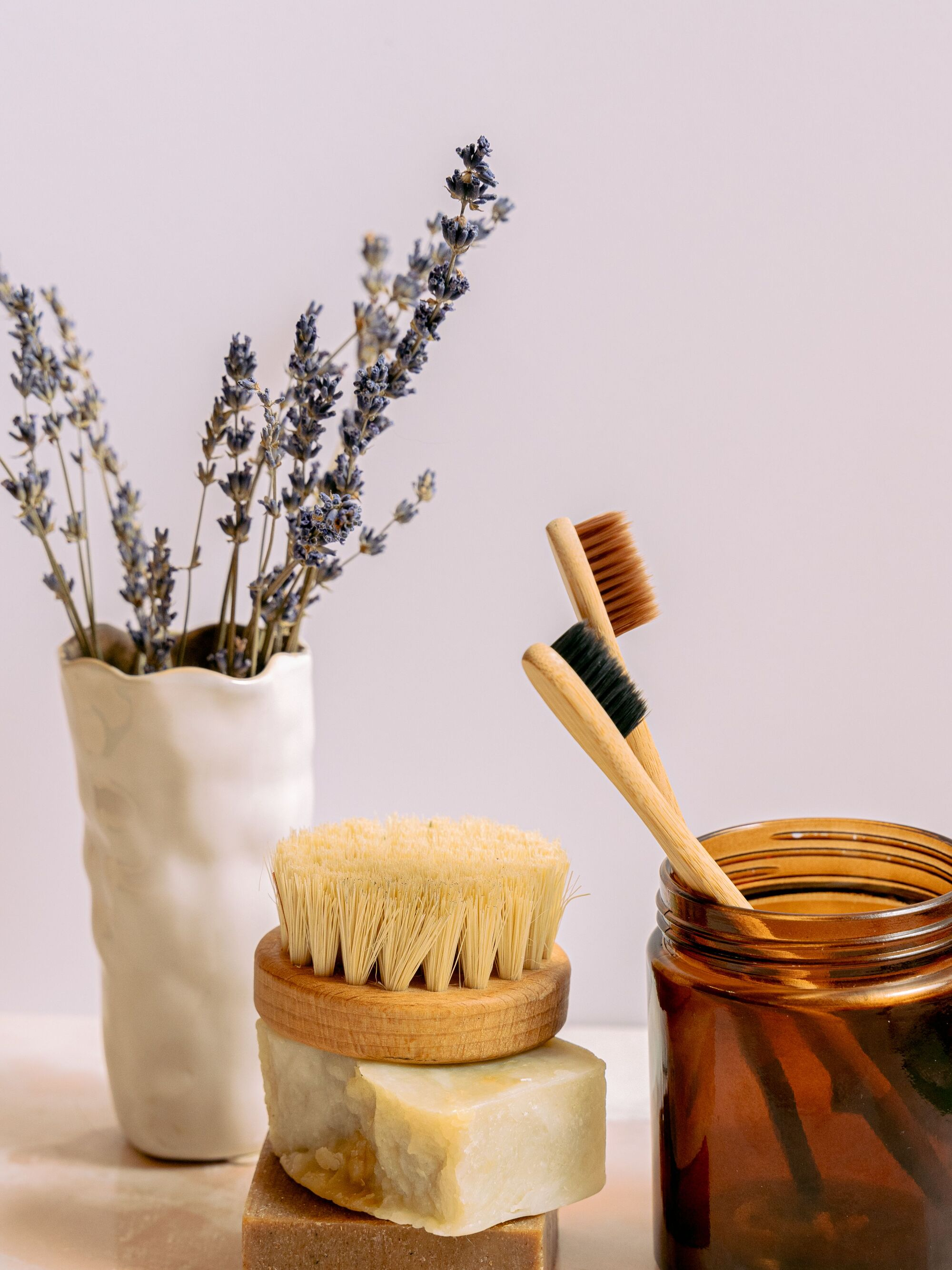 Dried lavender in a vase, stacked natural soaps, a dry brush, and two bamboo toothbrushes in an amber jar on a light surface.